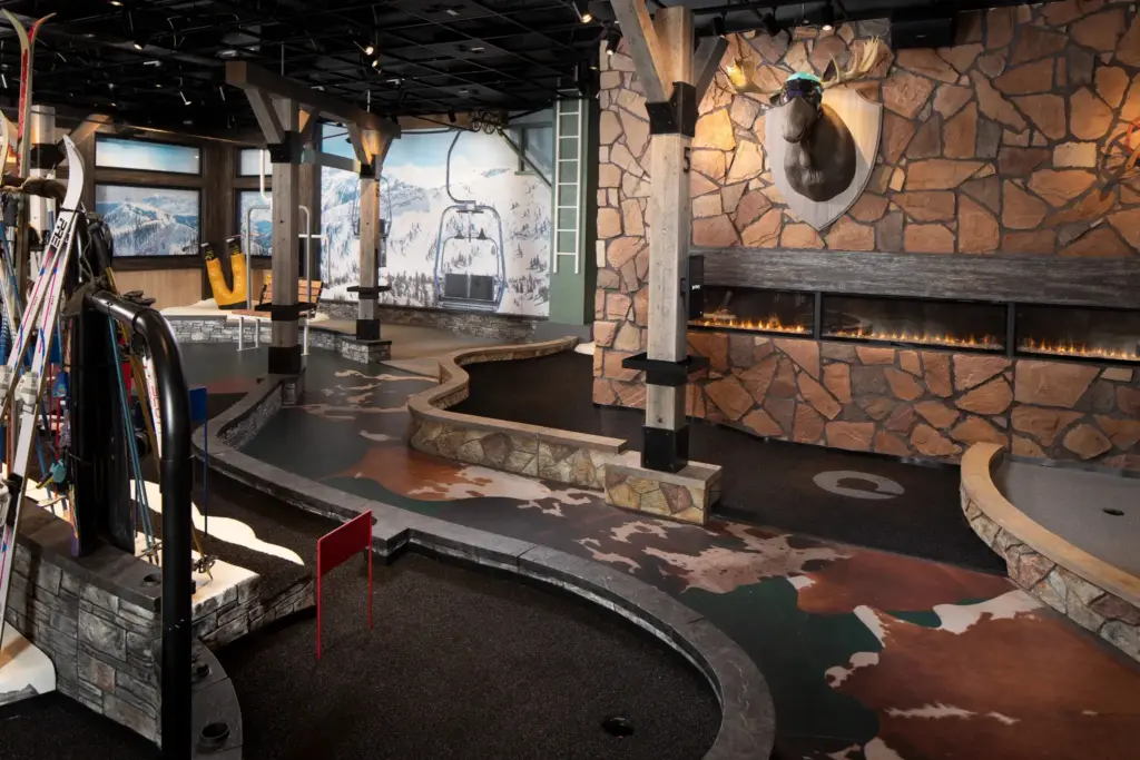 Lodge Course at Puttery Dallas featuring rustic stone and timber details, ski equipment, a mounted moose, and a fireplace beside winding putting lanes.
