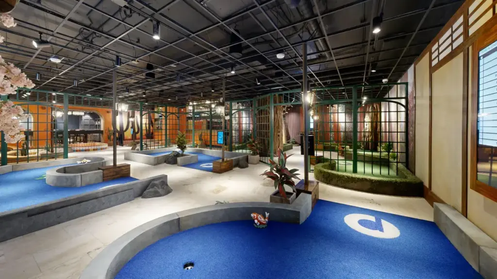 Wide view of the Conservatory Course at Puttery Kansas City with blue and green putting greens, garden trellises, plants and forest scenery.