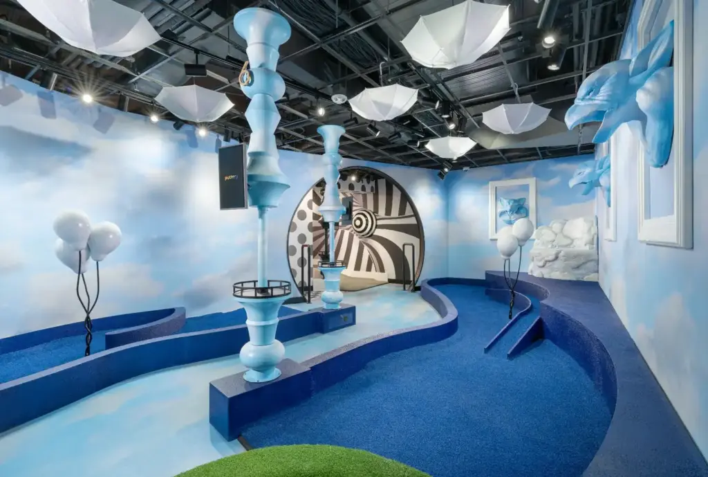 Dreamlike mini golf room with cloud-painted walls, hanging umbrellas, and blue course elements at the Washington DC Illusion course.