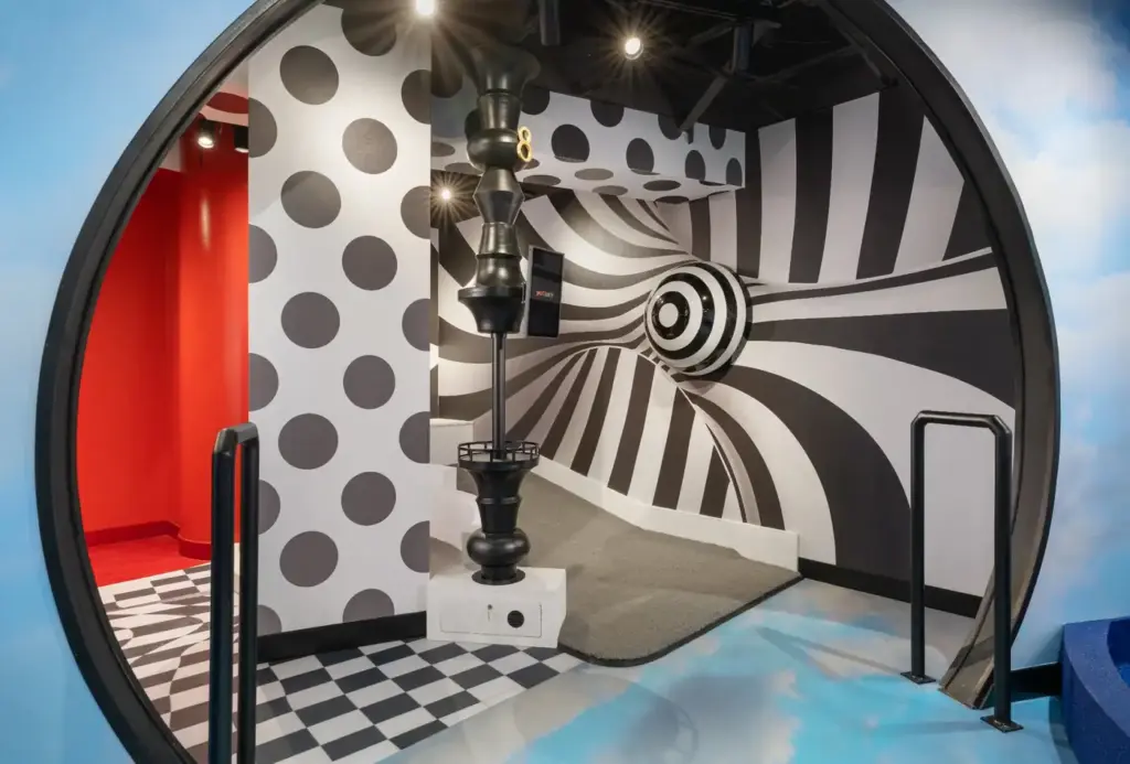 Entry view into a black-and-white illusion mini golf room with striped walls, polka dots, and a sculptural target feature at the Washington DC Illusion course.